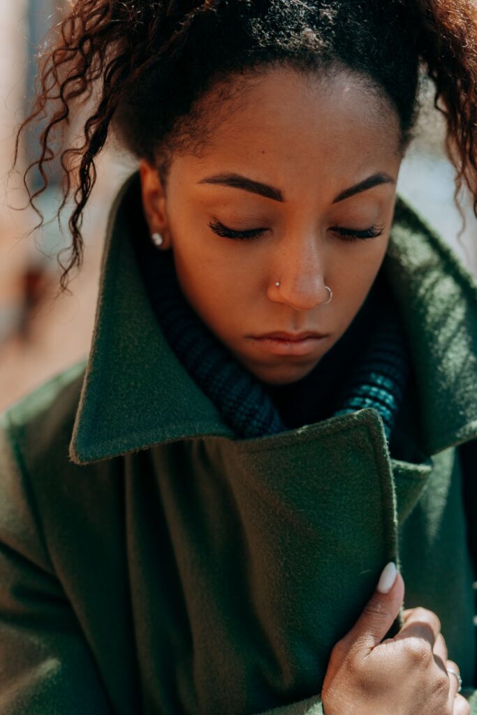 A thoughtful woman with curly hair and a nose ring wearing a green coat.