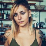 Close-up portrait of a tattooed young woman with blonde hair indoors, exuding confidence and style.