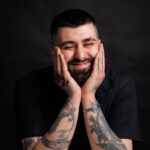 Portrait of a smiling man with tattoos leaning on a table in a studio.