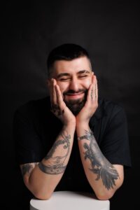 Portrait of a smiling man with tattoos leaning on a table in a studio.